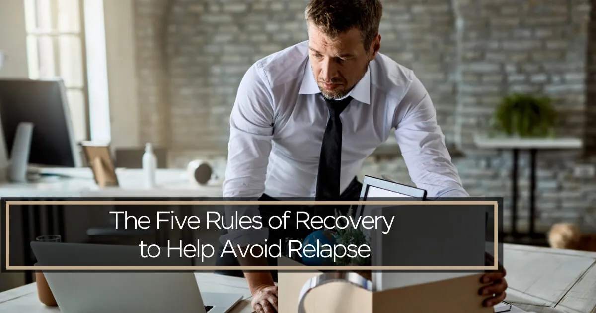 A man in a white shirt and black tie leans over a desk, looking down somberly at a cardboard box filled with office supplies.