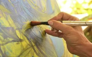 A hand using a brush to apply yellow paint to a blue abstract canvas.