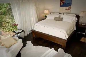 Modern bedroom with a dark wood bed, white linens, and a cowhide rug facing large windows with sheer curtains.