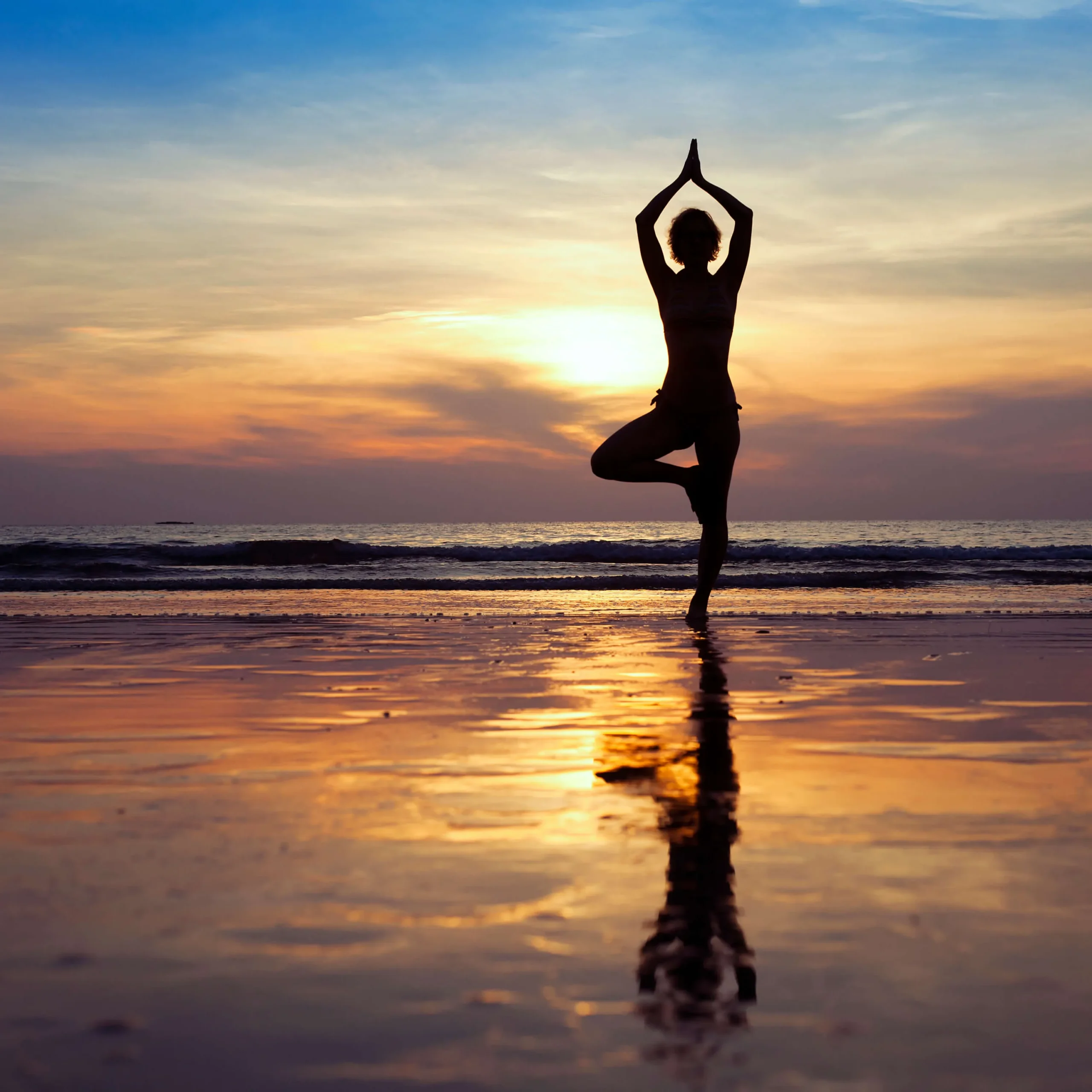 A silhouette of a person practicing the yoga tree pose on a beach at sunset.