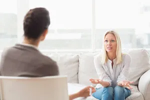 Blonde woman gesturing on a sofa while speaking to a therapist.
