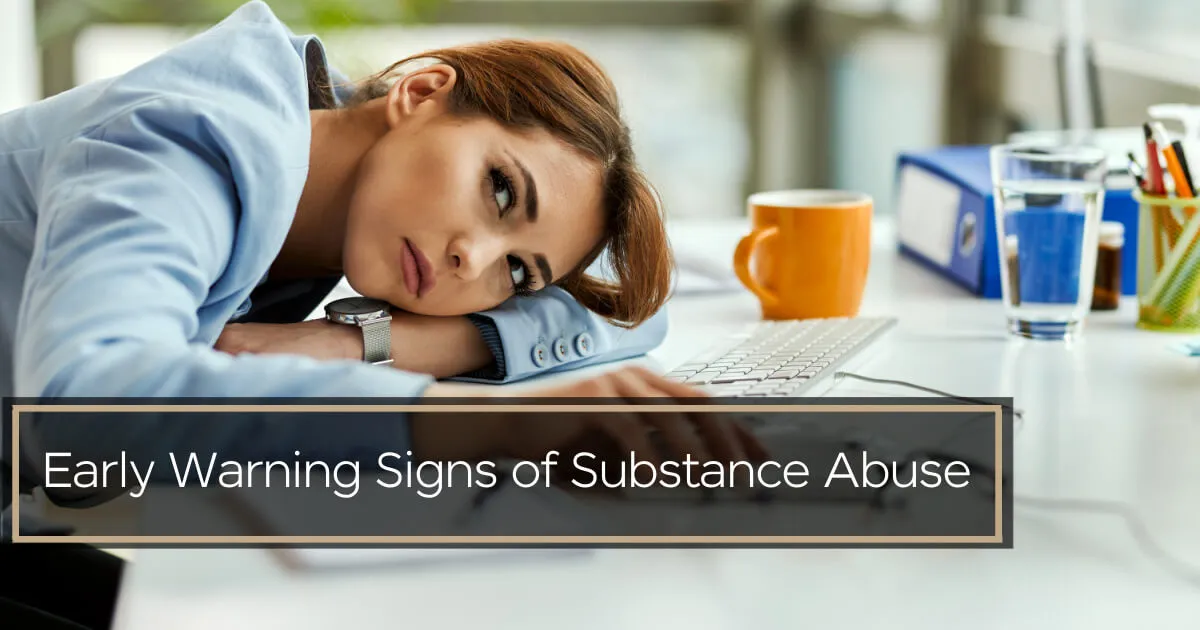 A woman in a blue blazer resting her head on an office desk, looking fatigued and despondent.
