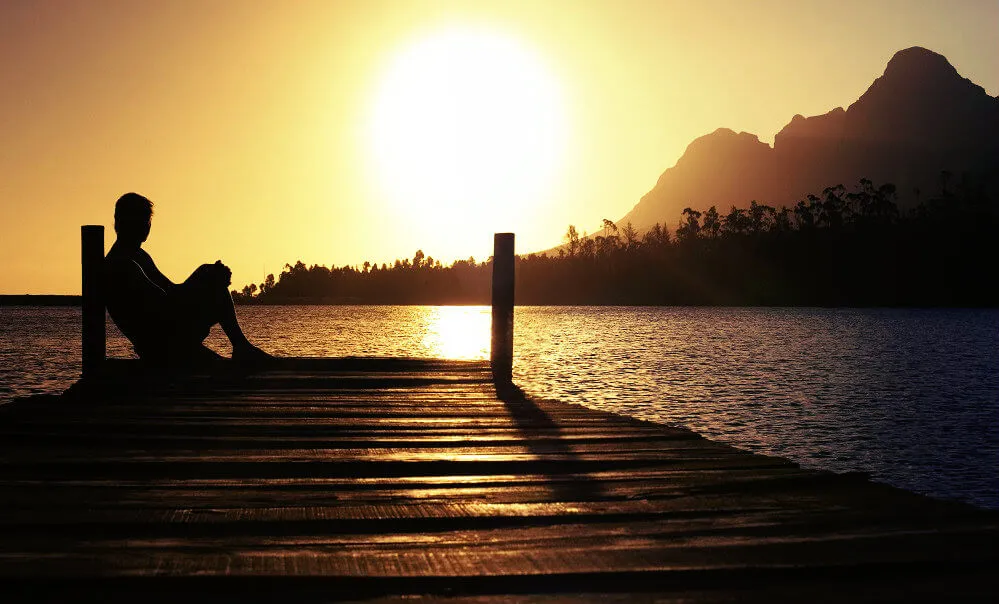 A silhouette of a person sitting on a wooden pier at sunset, looking out over a calm lake toward distant mountains.