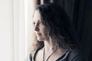 A thoughtful portrait of a woman with long, wavy brown hair looking out of a window.