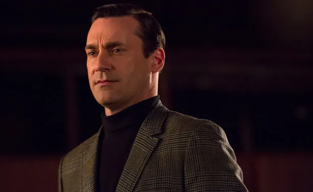 A man in a dark turtleneck and grey patterned blazer with a serious expression.