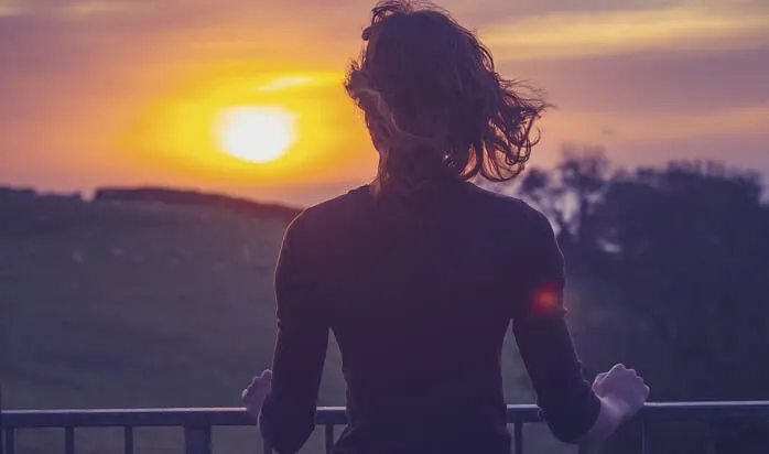 A woman watches the sunset over rolling hills from a balcony.