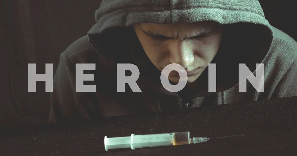 A person wearing a dark hoodie looks down at a filled syringe on a table.